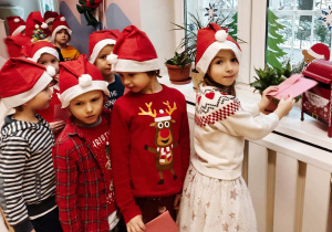 Dzieci stoją w rzędzie. Wrzucają do czerwonej skrzynki własnoręcznie przygotowane kartki do św Mikołaja.