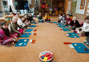 Dzieci siedzą w rzędach na podłodze. Przed sobą mają ułożone kolorowe figury geometryczne.