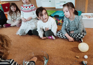 Dzieci siedzą na dywanie. Obok leżą: balon, korek, gwóźdź, moneta, piłka do ping ponga, gumowa zabawka.