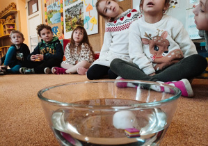 Dzieci siedzące na dywanie obserwują co się zadzieje z piłeczką do ping ponga, włożoną do miski z wodą.