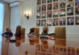 Dzieci siedzące przy stole w sali posiedzeń. Oglądają galerię zdjęć umieszczoną na jednej ze ścian.