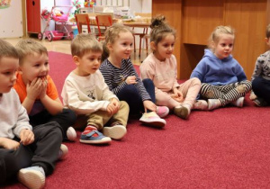 Dzieci siedzą na dywanie, słuchają prowadzącej zajęcia nauczycielki.