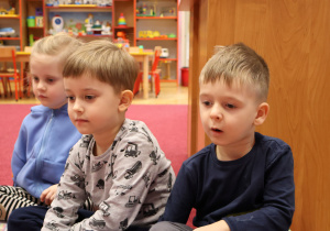 Dzieci siedzą na dywanie, słuchają prowadzącej zajęcia nauczycielki.