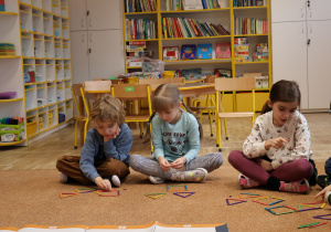 Dzieci siedzą na dywanie, układają z kolorowych patyczków figury geometryczne