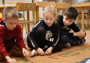 Dzieci siedzą na dywanie, układają z kolorowych patyczków figury geometryczne.