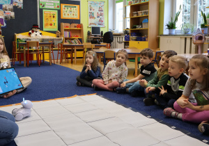 Dzieci siedza na dywanie, słuchają poleceń prowadzącej zajęcia.
