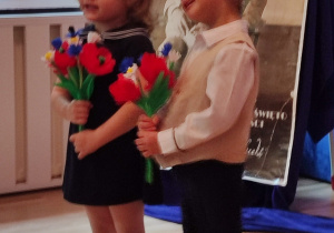 Dziewczynka i chłopiec trzymają bukiety kwiatów. Śpiewają piosenkę.
